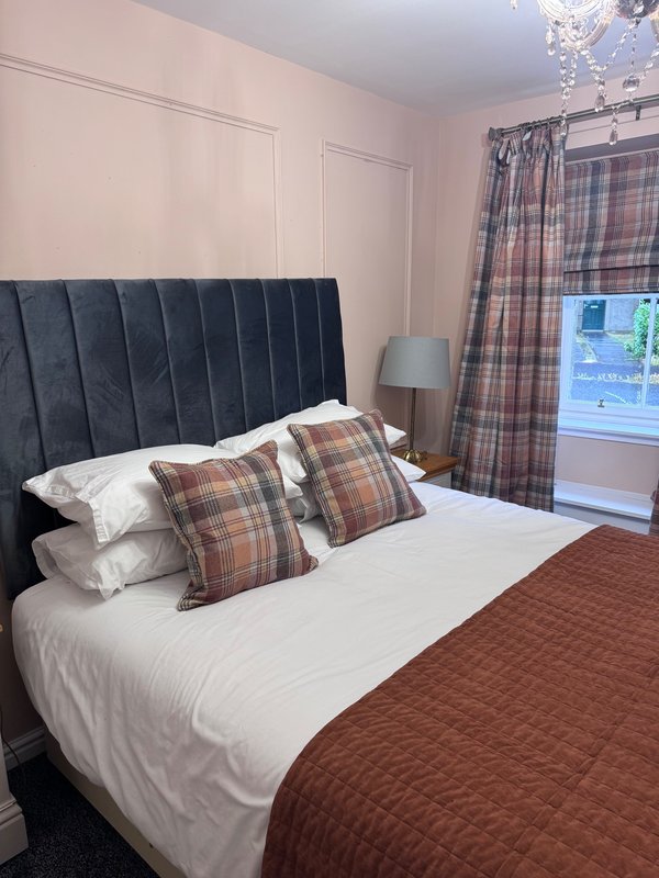 Bedroom with dark upholstered headboard, double bed, plaid cushions and tartan curtains