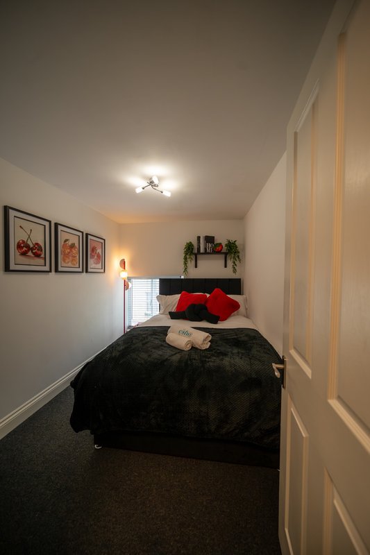 Compact bedroom with double bed, black bedding, red pillows, framed wall art and ceiling light