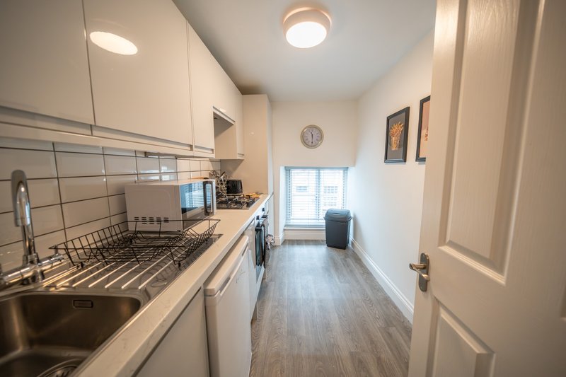 Bright galley kitchen with white cabinetry, stainless steel sink, tile backsplash and wooden flooring