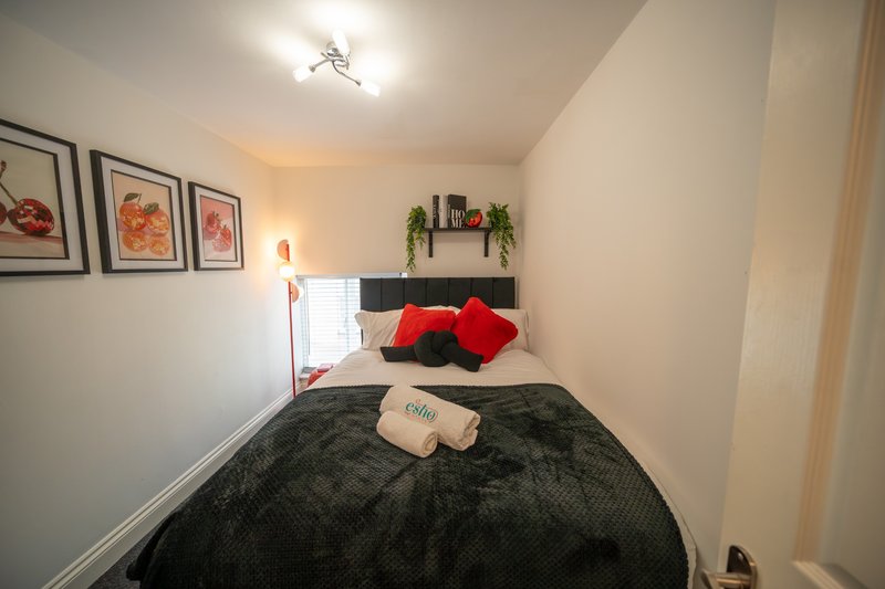 Small bedroom with single bed, dark bedding, red pillows, framed wall art, and potted plant