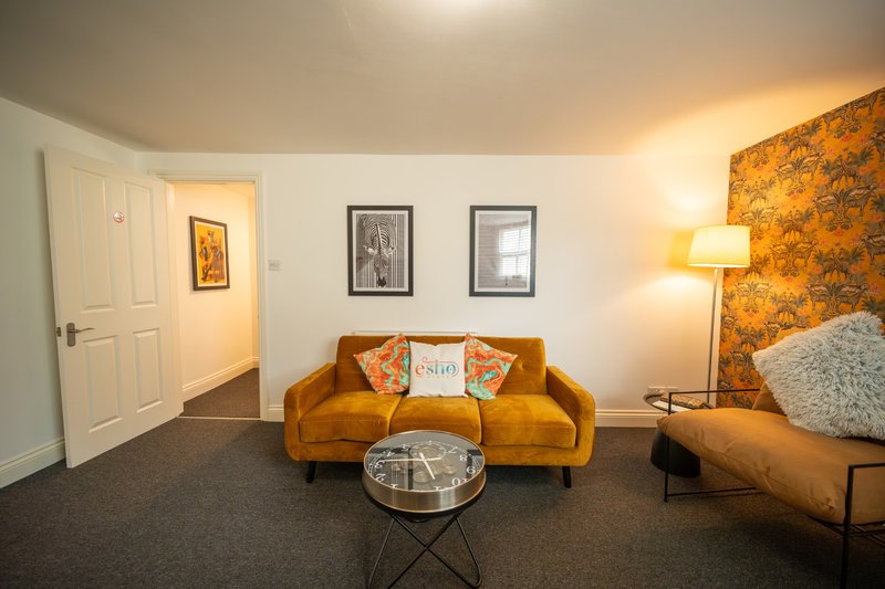 Living room with mustard-yellow sofa, framed artwork, side table lamp, and bed visible on right