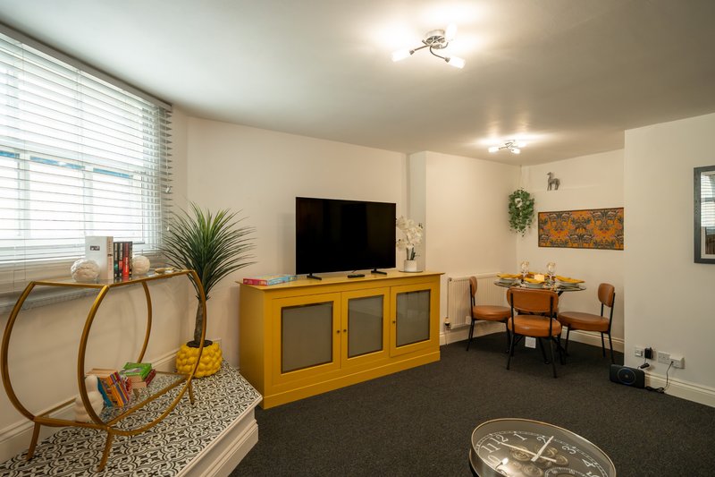 Living room with yellow TV unit, black TV, dining chairs, potted plant and window with blinds