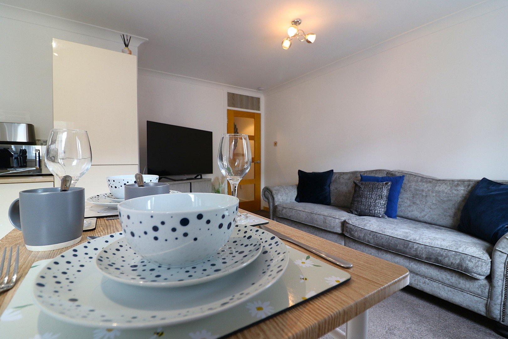 Open-plan living room with gray sofa, TV, kitchenette, and dining table with place settings