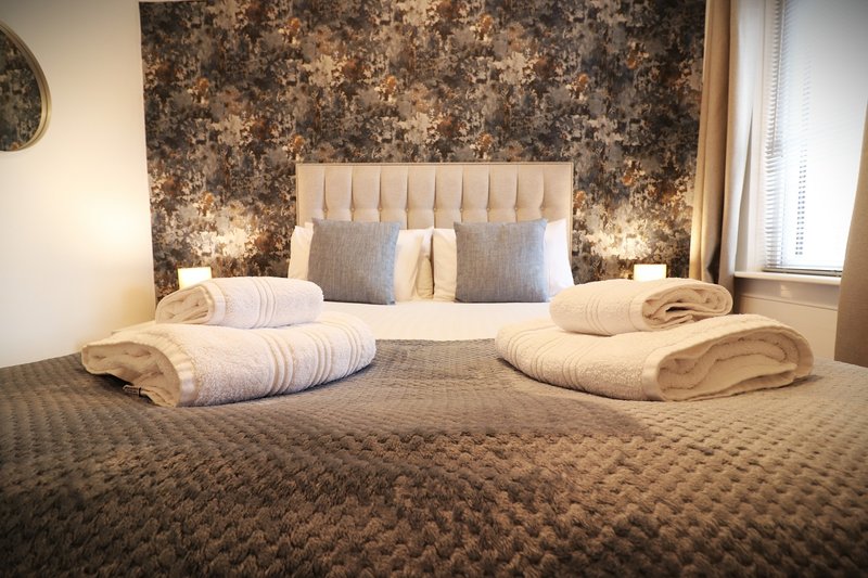 Bedroom with upholstered headboard, patterned wallpaper, rolled towels on bed, bedside lamps