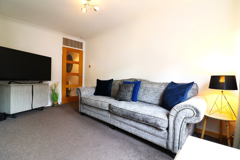 Living room with gray upholstered sofa, blue cushions, flat-screen TV, wooden shelving, and table lamp