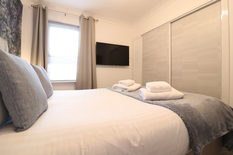 Modern bedroom with double bed, white bedding, cushions, wall-mounted TV, and window with curtains