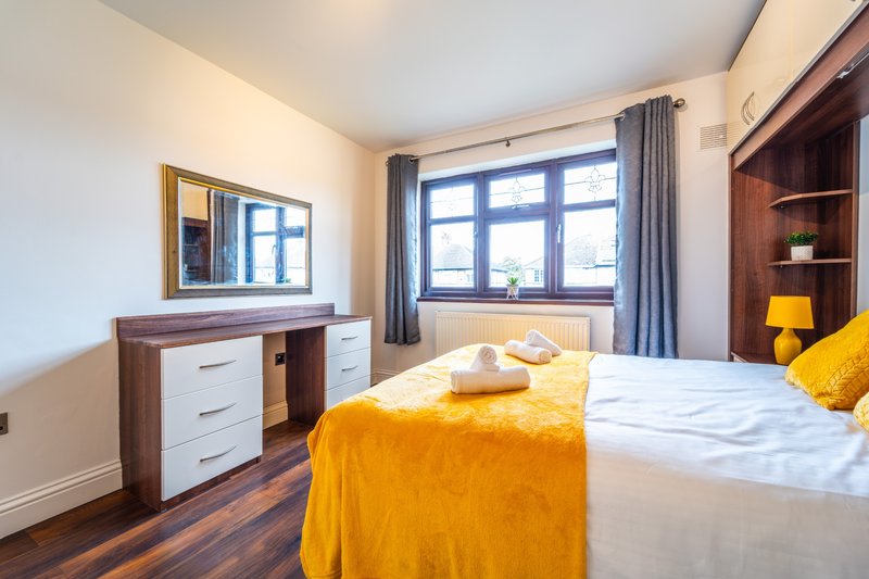 Bright bedroom with double bed, yellow throw, wooden dresser and large windows