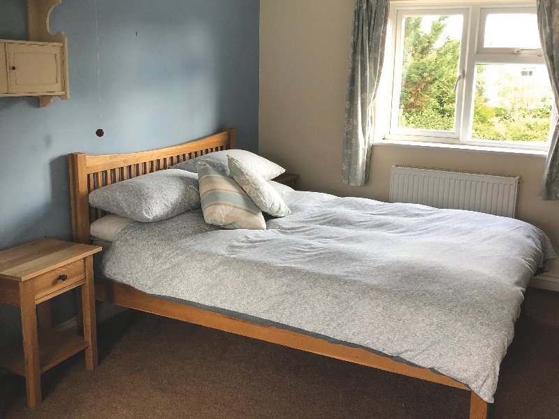 Bedroom with wooden bed, grey bedding, pillows, wooden nightstand and window with curtains