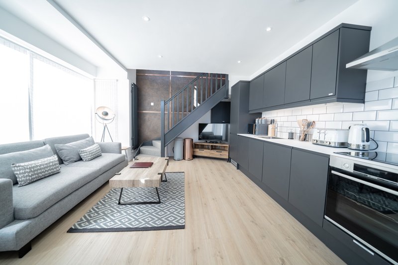 Open-plan studio with grey sofa, dark kitchen cabinetry, wooden flooring and mezzanine stairs