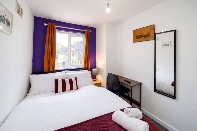 Compact bedroom with double bed, purple feature wall, window with gold curtains, wall mirror and small desk