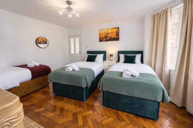 Bright bedroom with three single beds, green and burgundy bedding, wooden floor and cream curtains