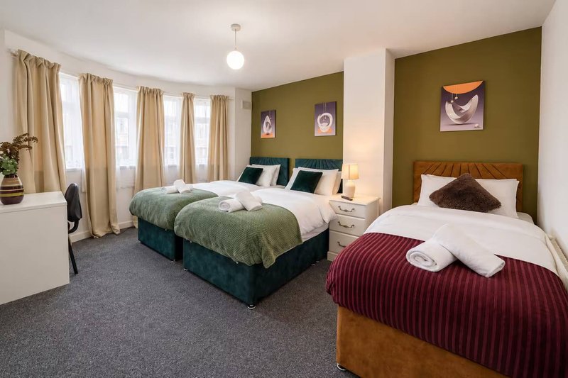 Spacious bedroom with two beds, olive-green accent wall, large windows with curtains, and bedside lamps