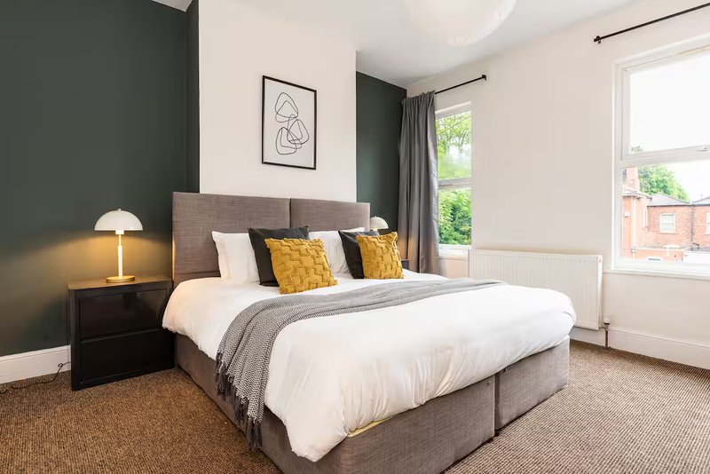 Bright bedroom with grey upholstered bed, white bedding, yellow cushions, dark accent wall, and window view