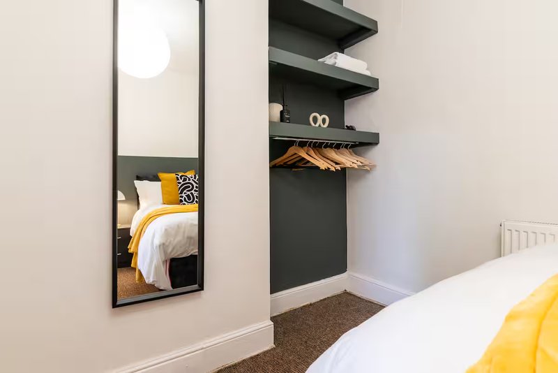 Modern bedroom with wall mirror, black floating shelves, yellow accent pillows and white bedding