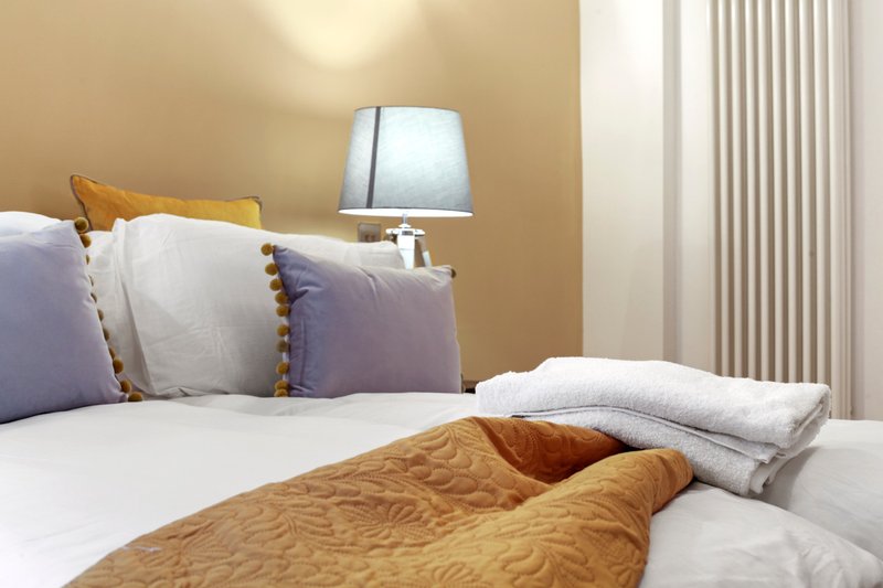 Bedroom with double bed, purple and gold cushions, bedside lamp and curtains