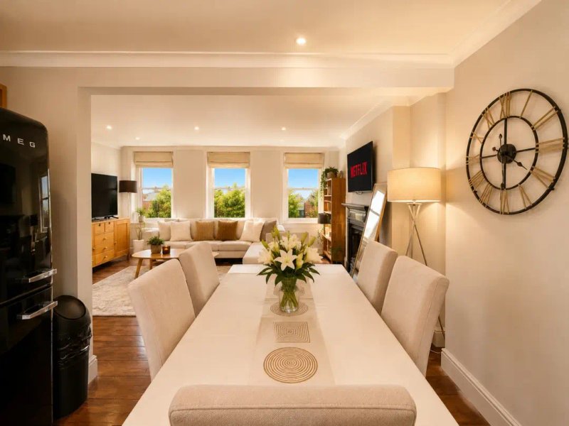 Open-plan dining and living room with white table, upholstered chairs, and cream sofa overlooking gardens through large windows