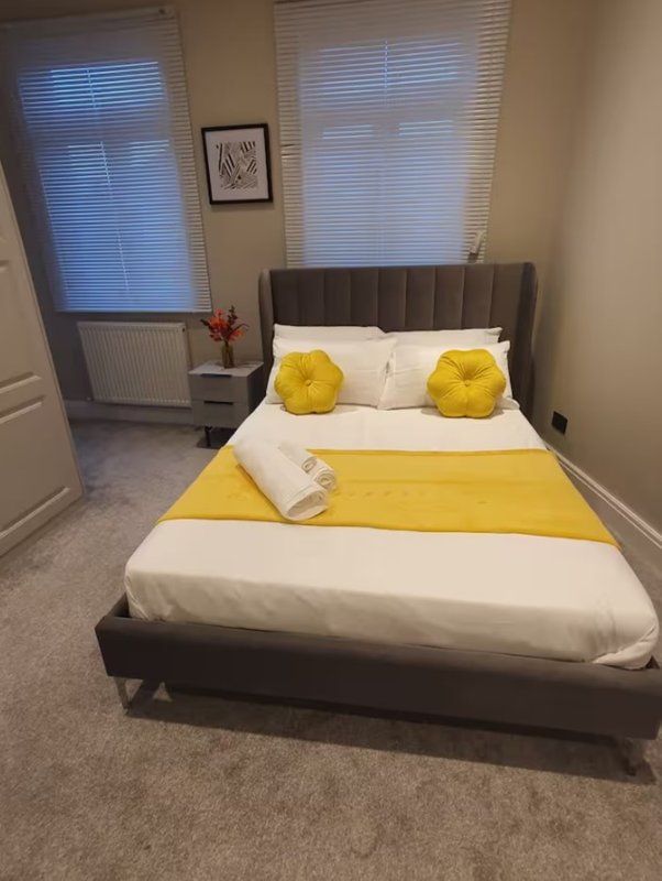 Bedroom with double bed, yellow and white bedding, two windows with blinds, and framed wall art