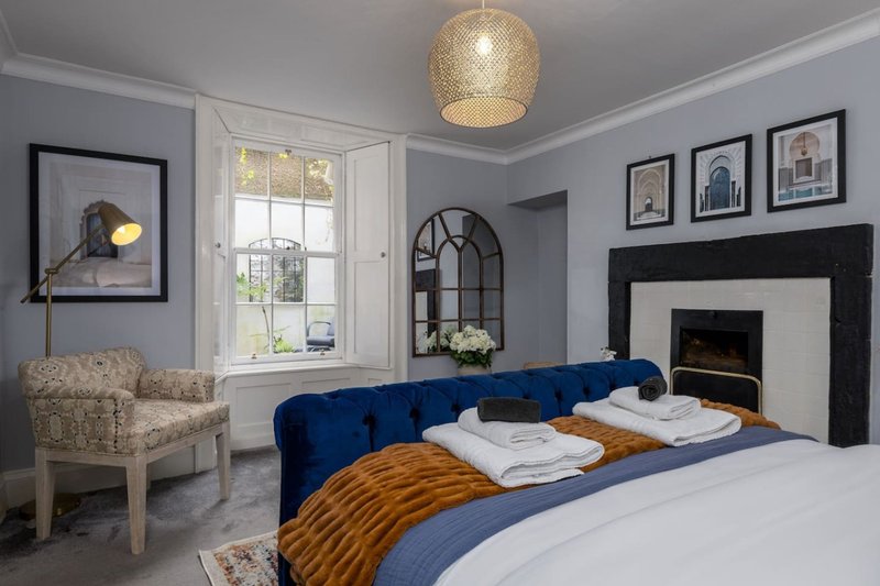 Bedroom with blue velvet headboard, fireplace, cream armchair and framed wall art