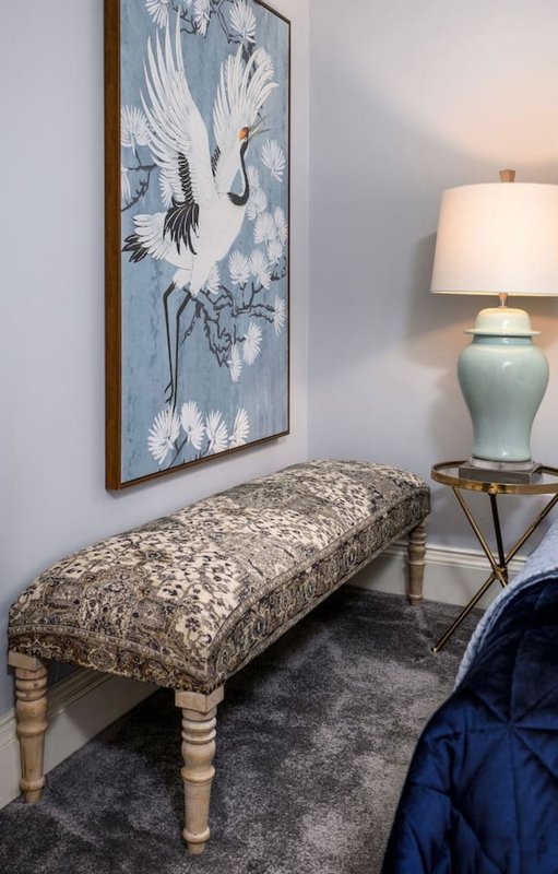 Bedroom corner with upholstered bench, framed crane artwork, table lamp and rug