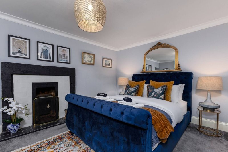 Bedroom with blue upholstered bed, fireplace, framed wall art, gold mirror and bedside lamps