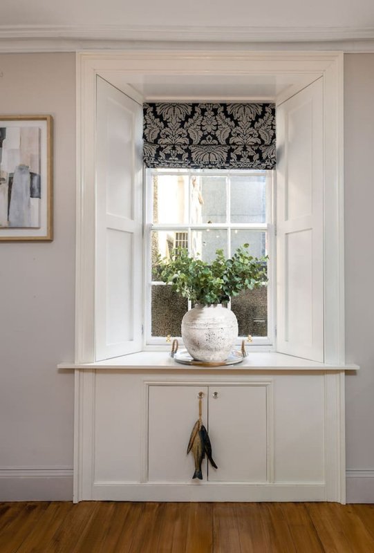 Bright window seat with white cabinetry, decorative plants, and patterned roman blind