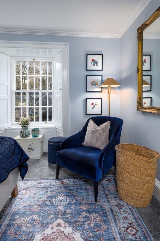 Bedroom corner with blue velvet armchair, framed botanical prints, table lamp and wicker basket