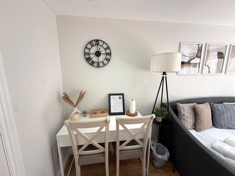 Bedroom corner with desk, two chairs, wall clock, lamp and bed with cushions