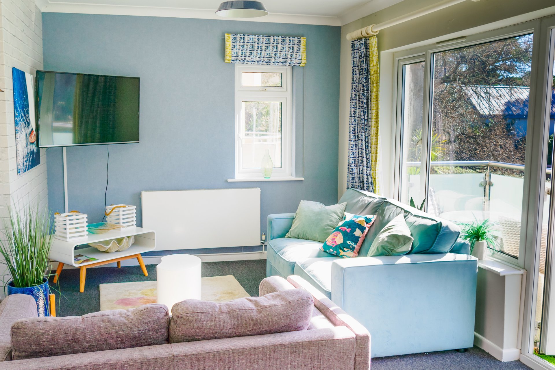 Bright living room with blue walls, sofa, desk, TV, and garden views through glass doors