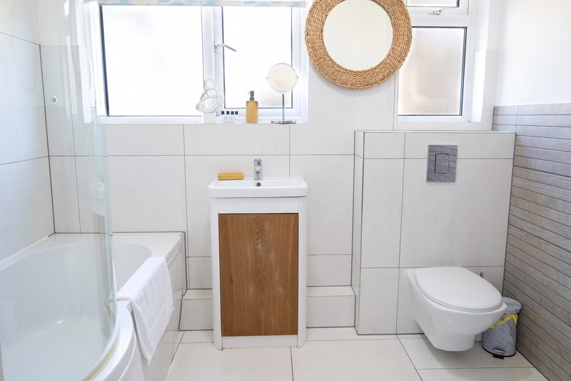 Clean bathroom with white tiles, wooden vanity sink, toilet, bathtub and round mirror.
