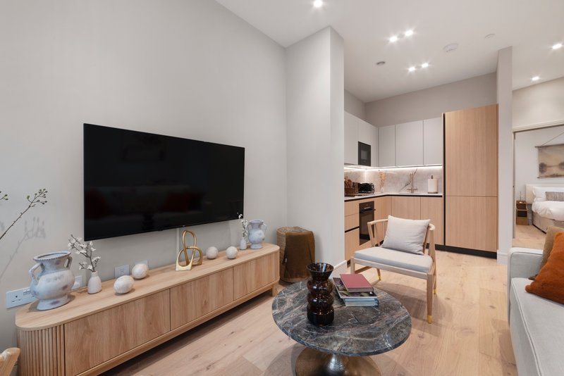 Modern living room with wall-mounted TV, wooden media console, armchair and open-plan bedroom visible