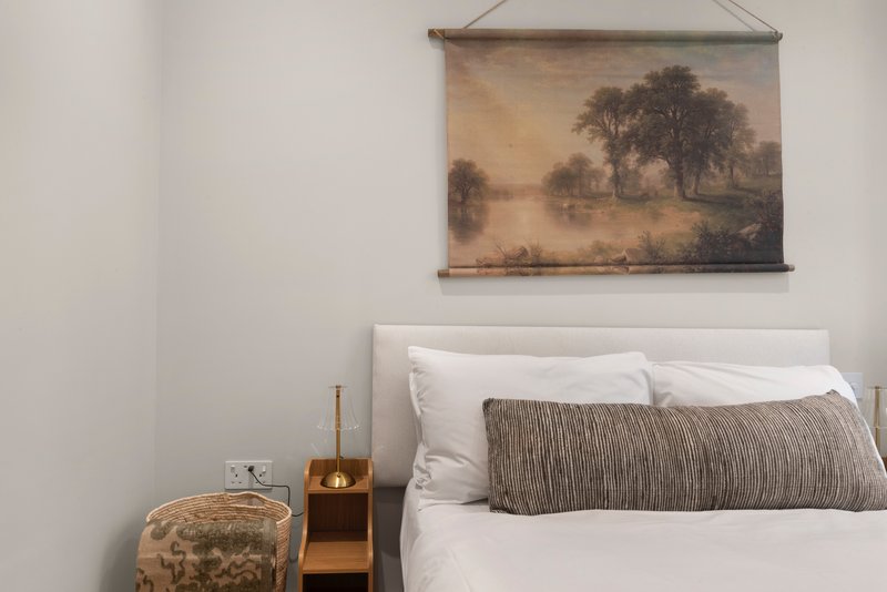 Bedroom with white bedding, grey cushion, wooden nightstand, and landscape wall art