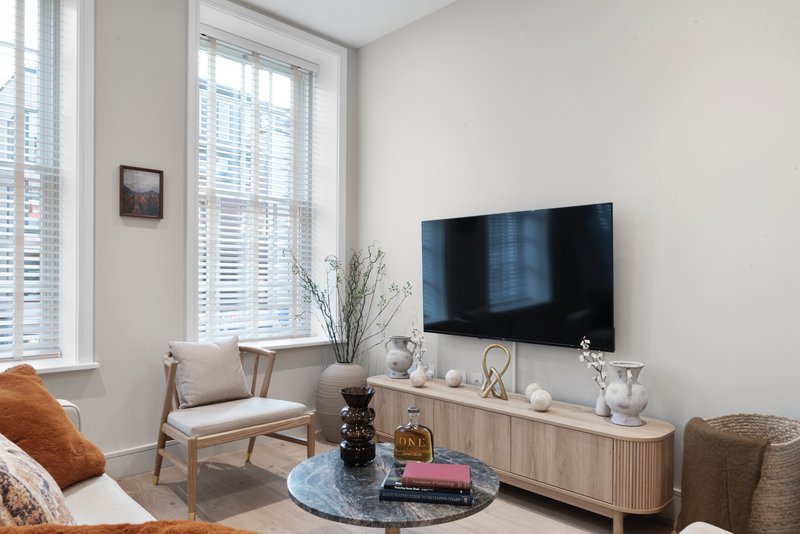 Modern living room with wall-mounted TV, wooden media console, armchair, and large windows with shutters