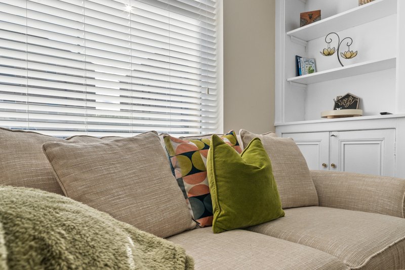 Neutral living room with beige sofa, decorative cushions, white shelving, and venetian blinds