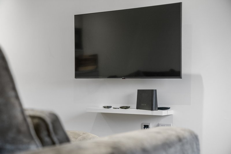 Wall-mounted flat-screen TV above white shelf with speaker and remote controls