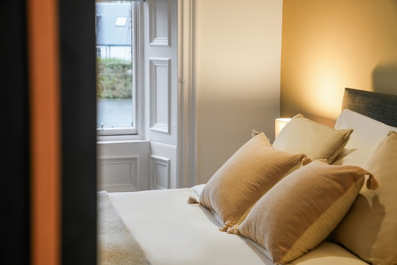 Bright bedroom with double bed, gold cushions, white bedding, and view through open door to window