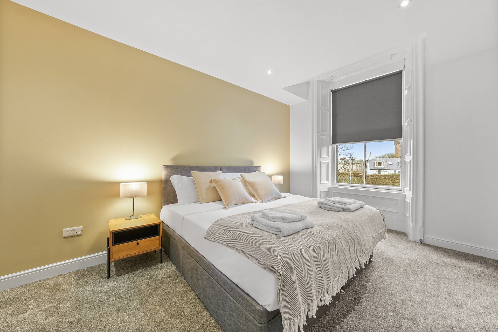 Bedroom with mustard-yellow accent wall, double bed, bedside table with lamp, and window
