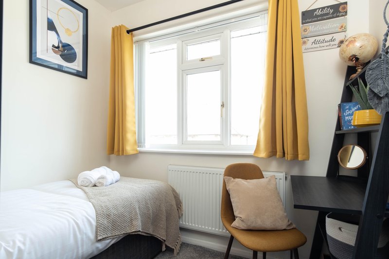 Small bedroom with single bed, yellow curtains, wooden chair, desks area and window with radiator