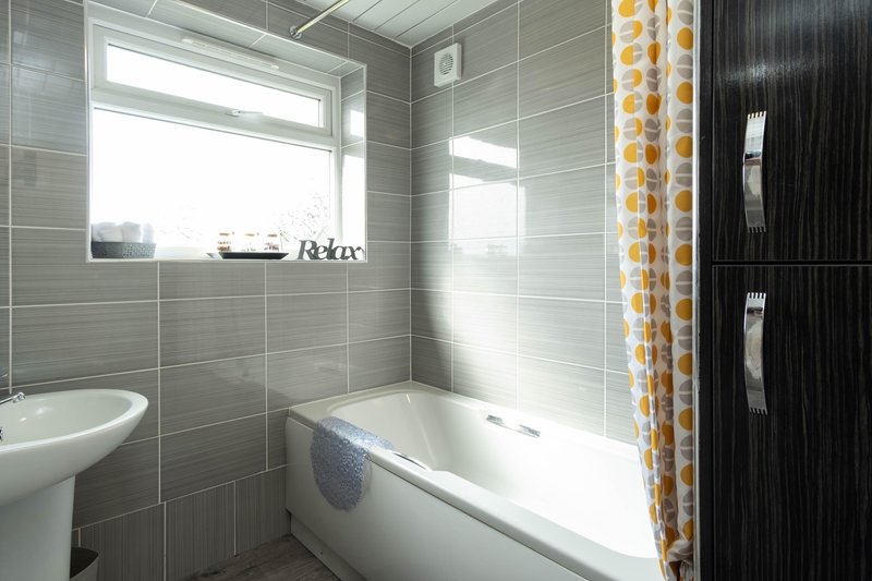 Modern bathroom with white bathtub with overhead shower, pedestal sink, gray tile walls, and yellow patterned shower curtain