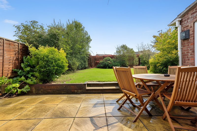 Patio area with wooden dining table and chairs overlooking spacious garden with lawn and trees