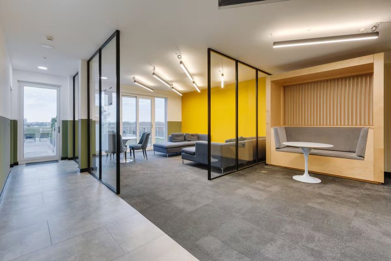 Modern open-plan office space with glass partitions, yellow accent wall, and lounge seating area