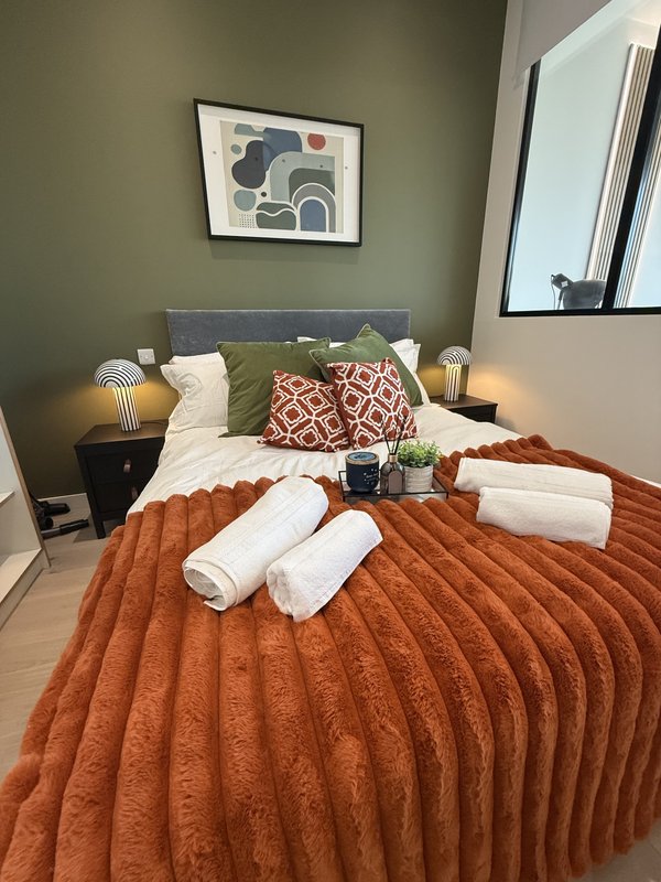 Bedroom with double bed, orange bedding, green headboard, rolled towels, and framed wall art