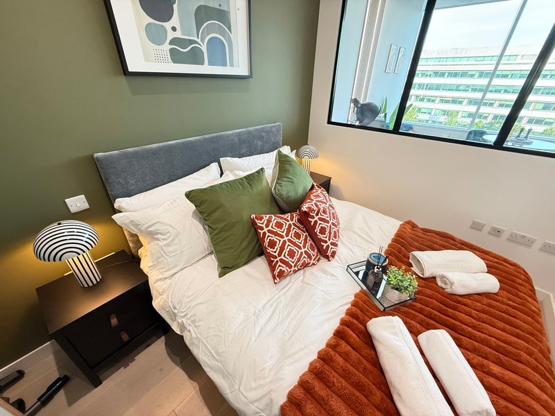 Modern bedroom with double bed, grey headboard, colourful cushions, bedside lamp and city views