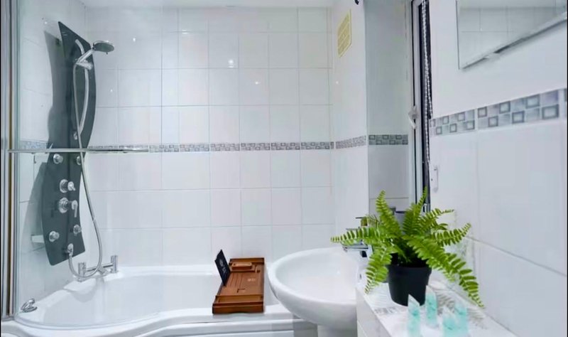 Modern bathroom with white tile walls, bathtub, toilet, shower head, and potted fern plant on shelf