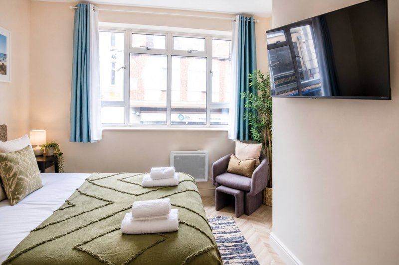 Bright bedroom with double bed, armchair, wall-mounted TV and large window with blue curtains