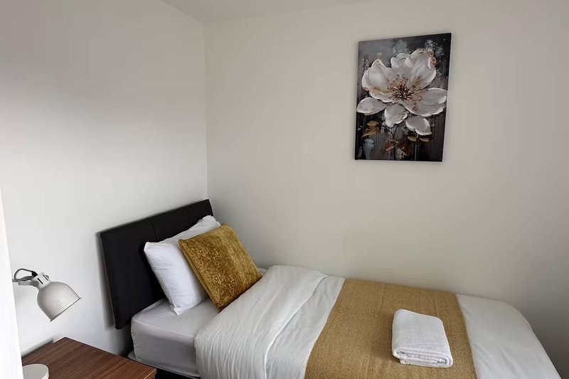 Bedroom with single bed, white bedding, gold cushion, wall-mounted flower artwork and bedside lamp