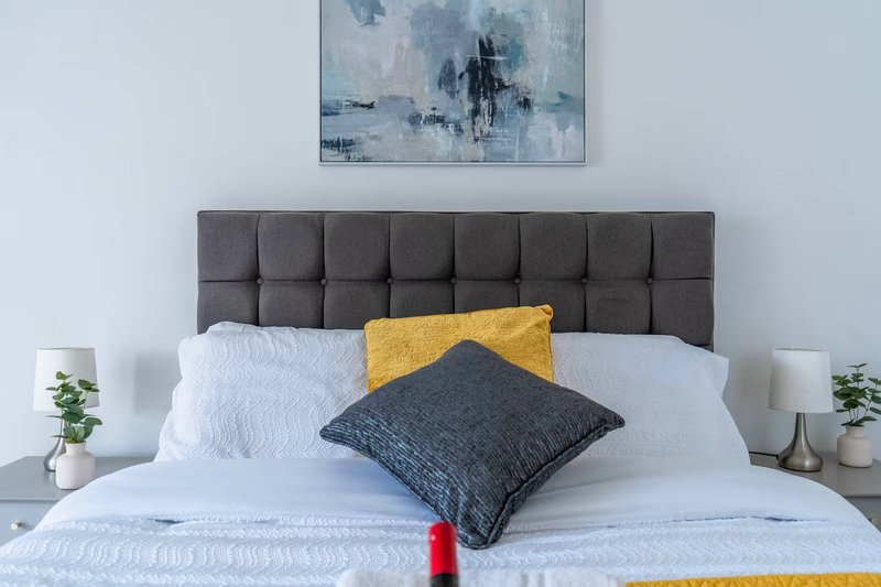 Bedroom with gray upholstered headboard, white bedding, yellow and dark pillows, and wall art above
