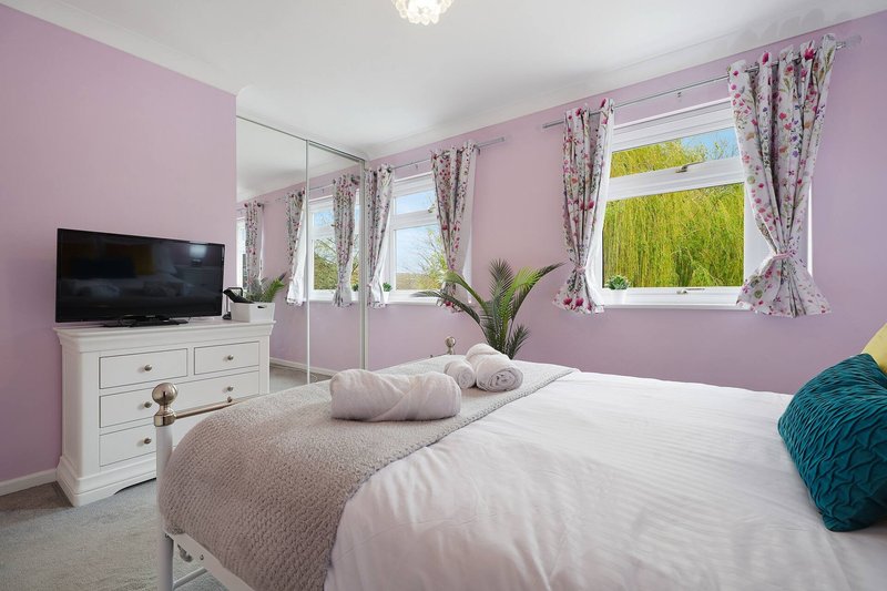 Bright bedroom with pink walls, double bed, white dresser, TV, and large windows with garden views