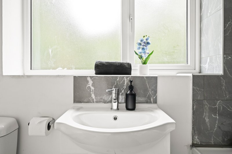 Clean bathroom with white pedestal sink, grey marble backsplash, window and toiletries