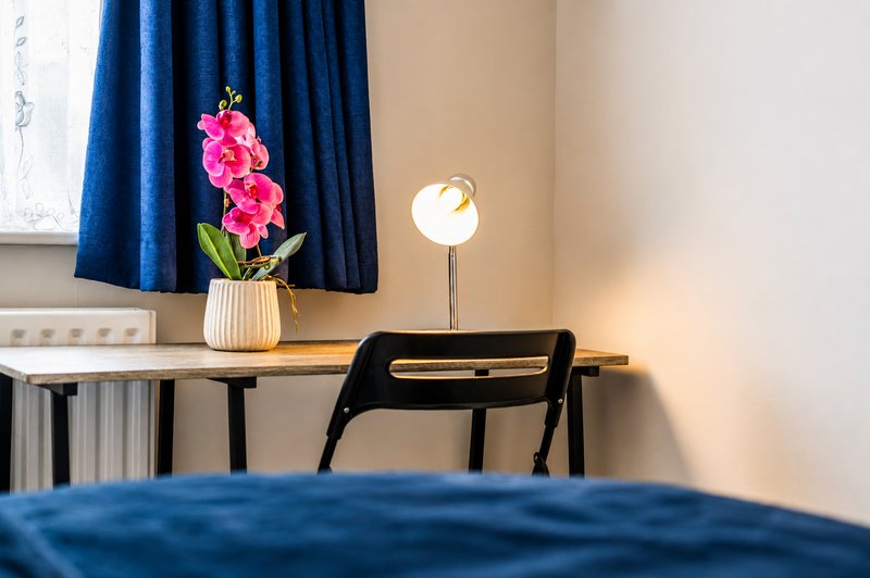Bedroom desk with pink flowers, lamp, and black chair near blue curtains