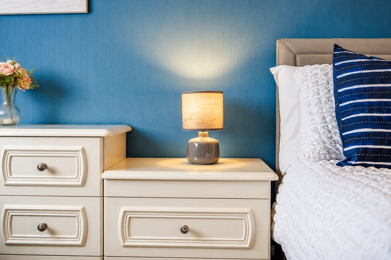 Bedroom with blue walls, white nightstand, bedside lamp, and bed with navy pillows